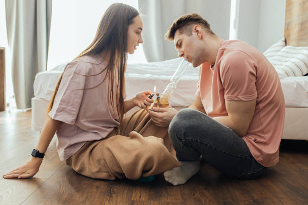 caucasian couple sit on the floor in the bedroom, woman help to light a bong bowl with cannabis. they use marijuana, weed. legalization of cannabis
